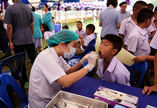 Dr. Waranuch Pitiphat, dressed in a white lab coat, provides dental care to a child. They are pictured seated within a tent where other dentists work with other patients. 