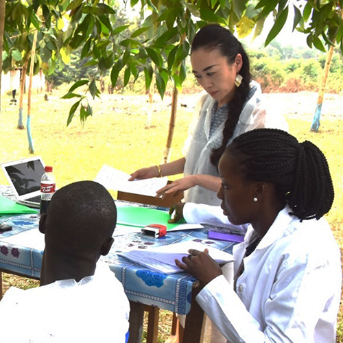 This photo shows Rie Yotsu (standing, right) reviewing paper work with mentees (seated). They are around a table set up outdoors